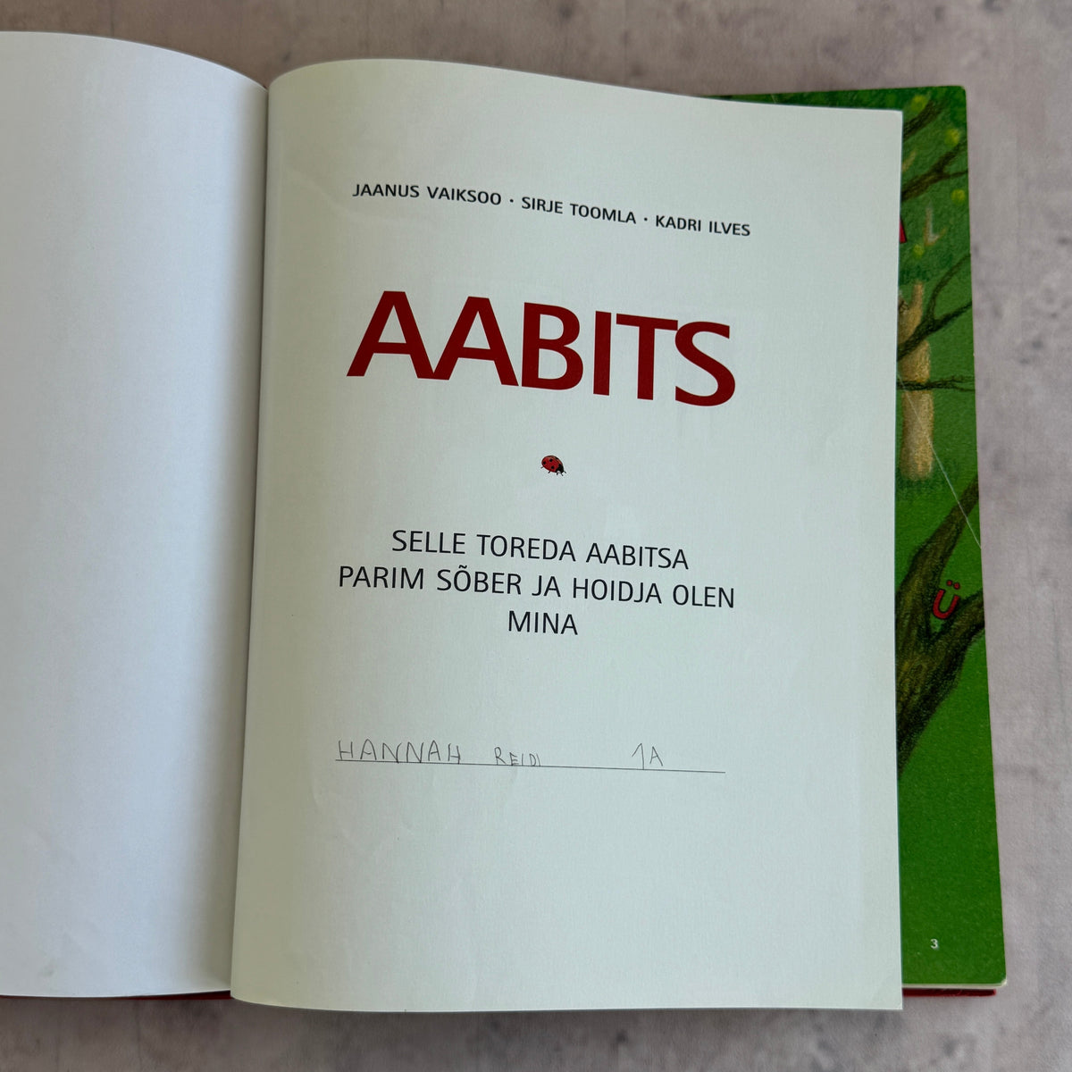 An open book showing the title page of 'AABITS' in large red font on an off-white page, with a green illustrated cover partially visible underneath.